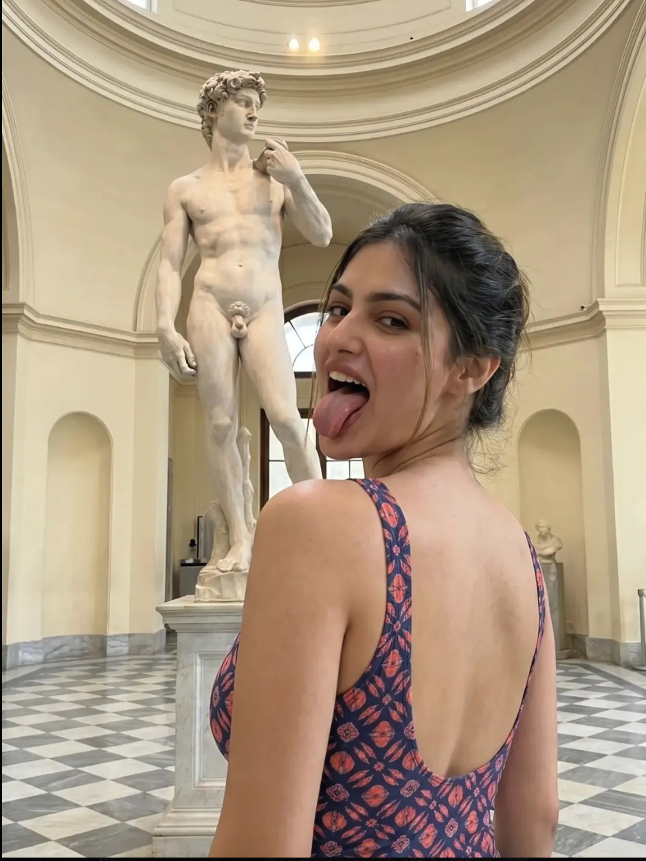 A woman looks over her shoulder and sticks her tongue out playfully while posing in a museum gallery in front of Michelangelo's marble statue of David.