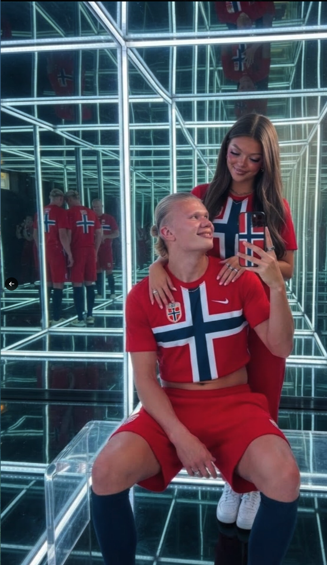 Erling Haaland and a woman posing for a mirror selfie in matching red Norway national team gear inside a brightly lit infinity mirror cube room.