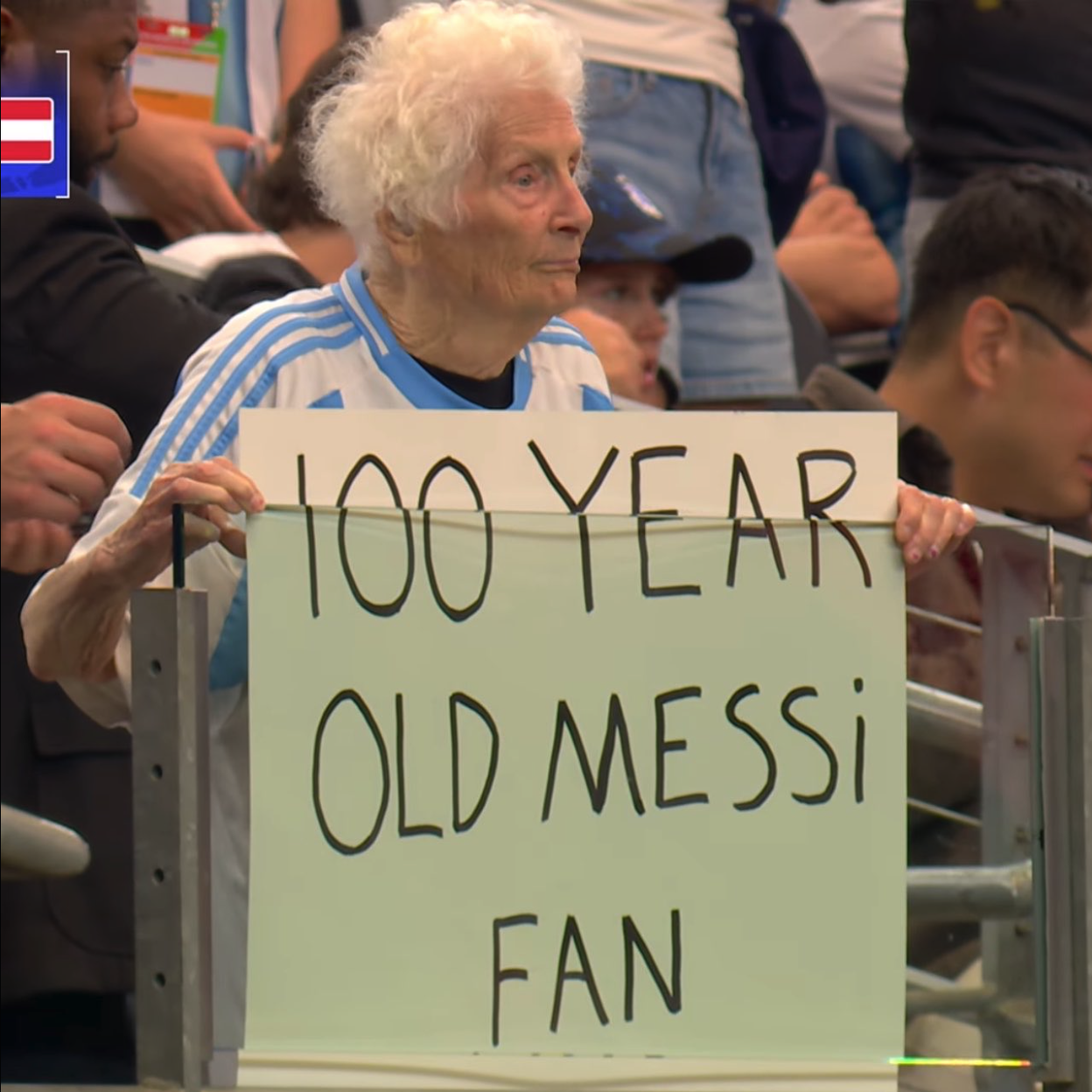 An elderly person holding a handwritten '100 year old Messi fan' sign in stadium stands.