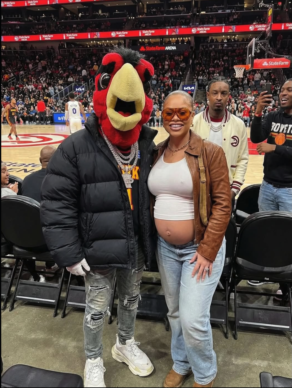 A person in a red bird mascot costume resembling 21 Savage posing with a pregnant woman courtside at a basketball arena.
