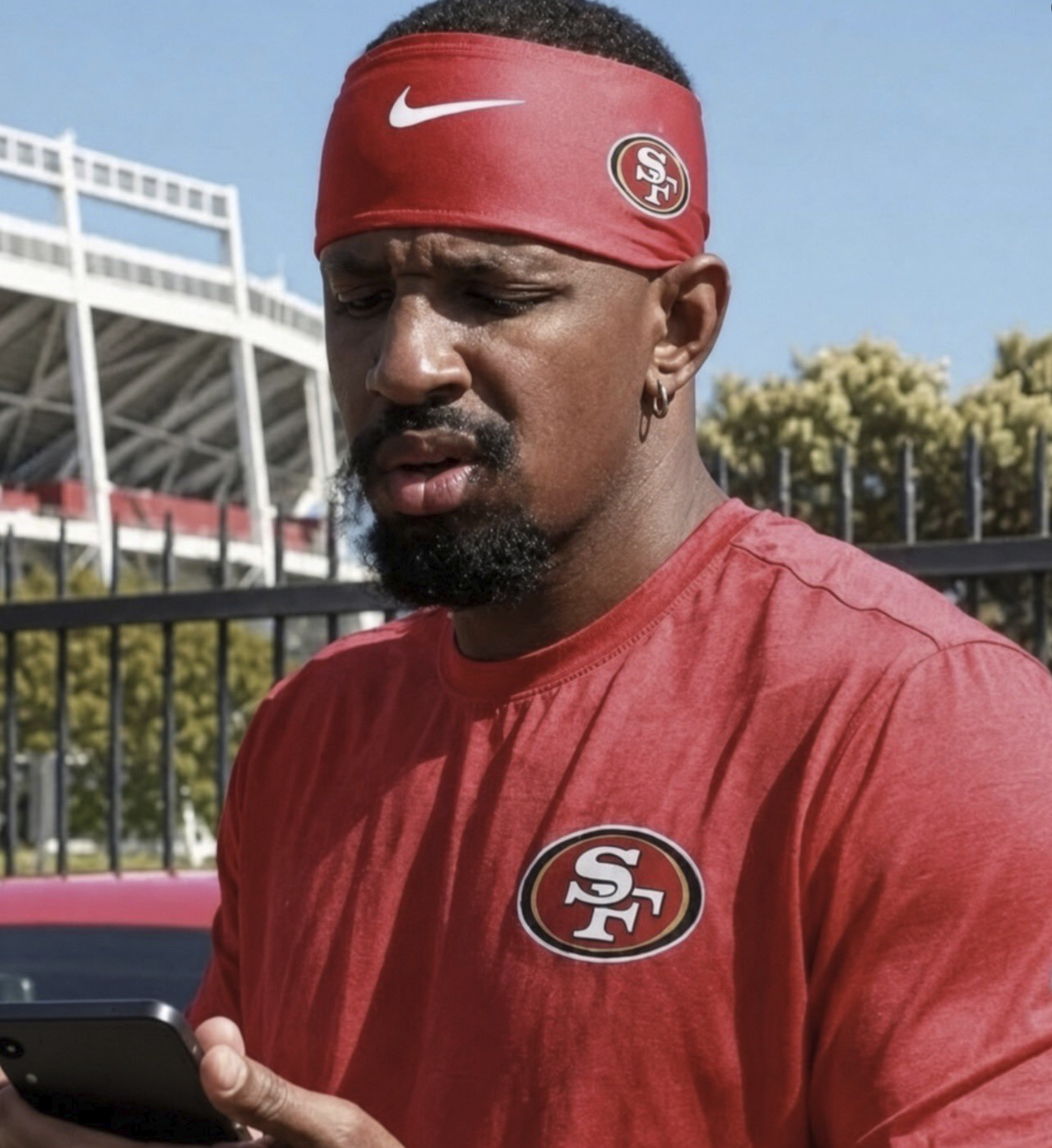 A man in a red San Francisco 49ers shirt and matching headband holding a smartphone outside a stadium.