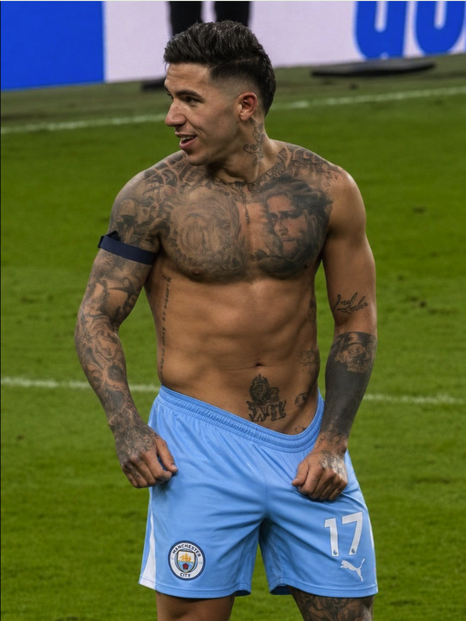 A shirtless, tattooed man resembling Adrián Ugarriza standing on a grass pitch in light blue Manchester City shorts.