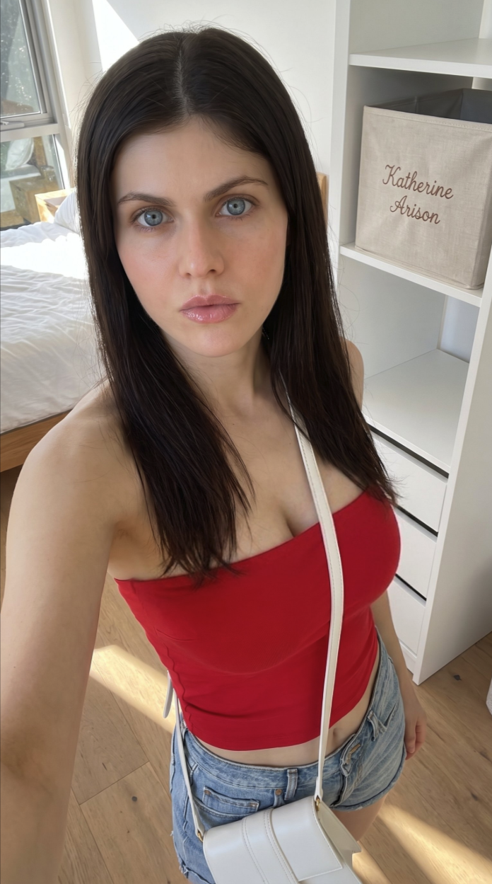 A woman resembling Alexandra Daddario wearing a red tube top and denim shorts, taking a selfie in a bedroom.