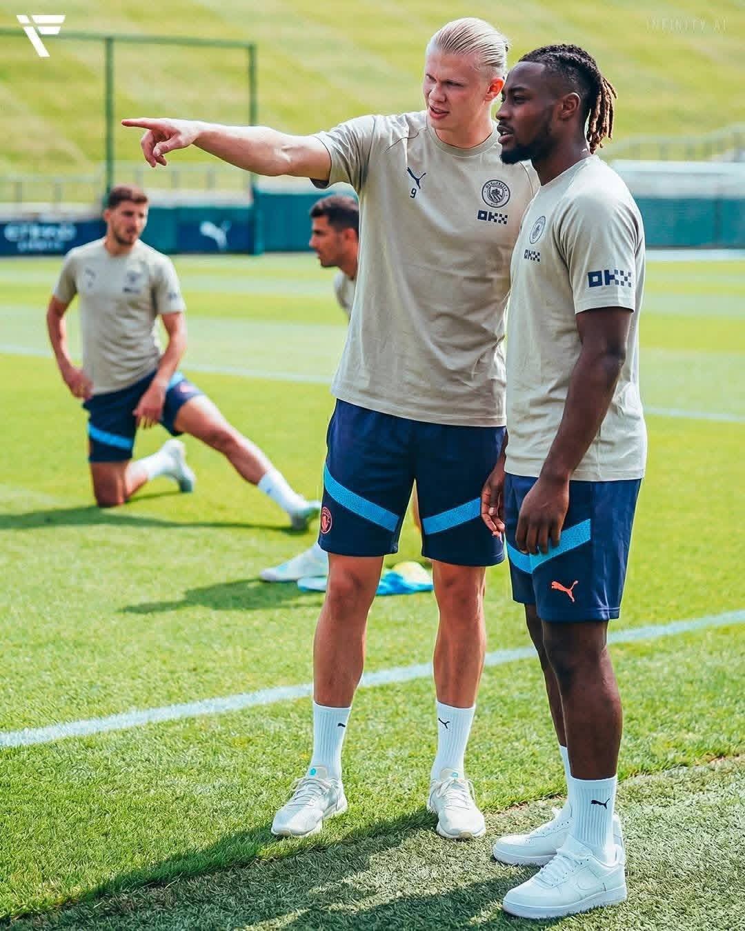 Two soccer players in Manchester City training gear on a grass pitch, one resembling Antonio Semenyo.