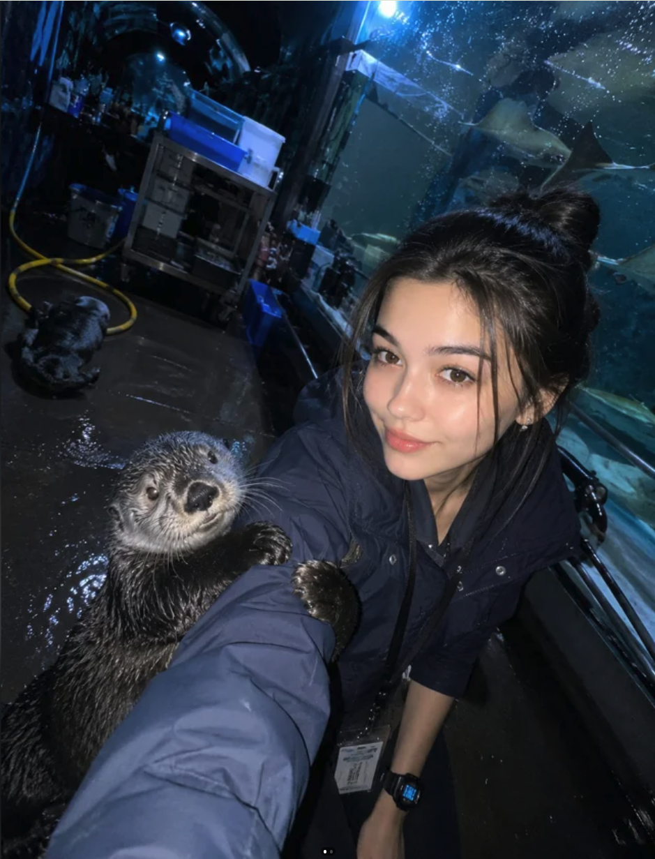 A young woman with dark hair taking a selfie with a wet otter in an aquarium back-of-house area.