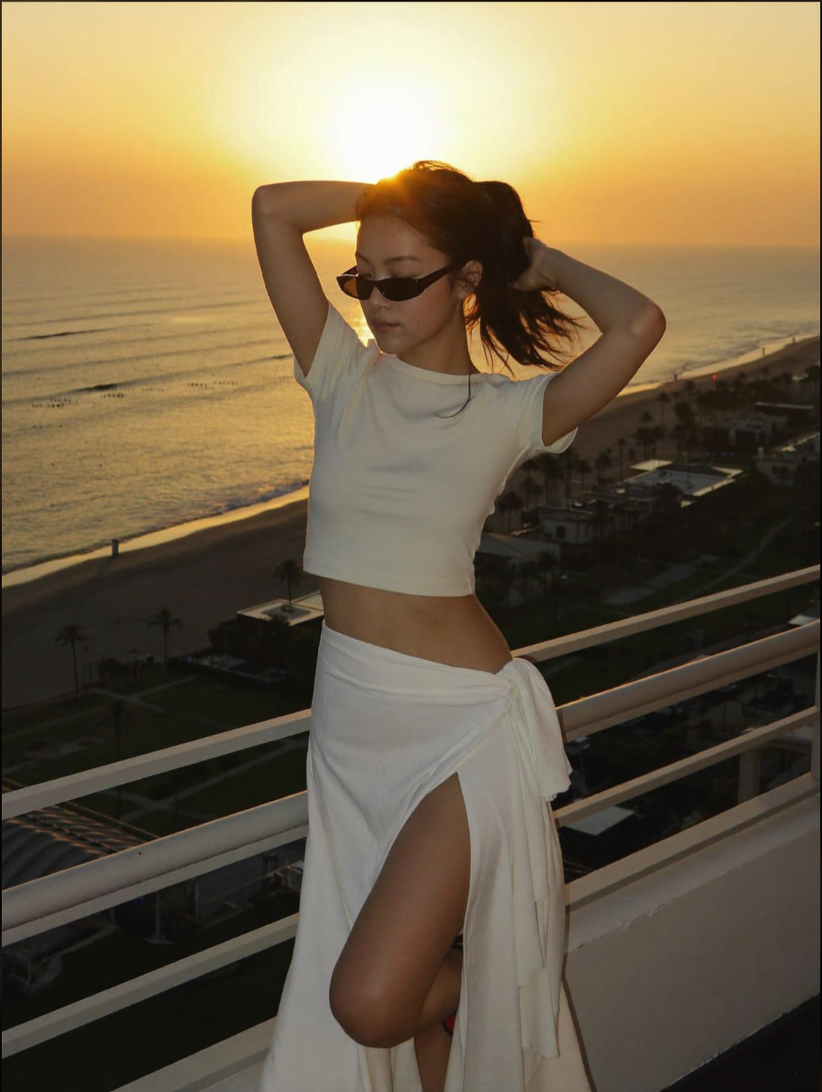 A woman in a white crop top and wrap skirt standing on a balcony at sunset with the ocean behind her.