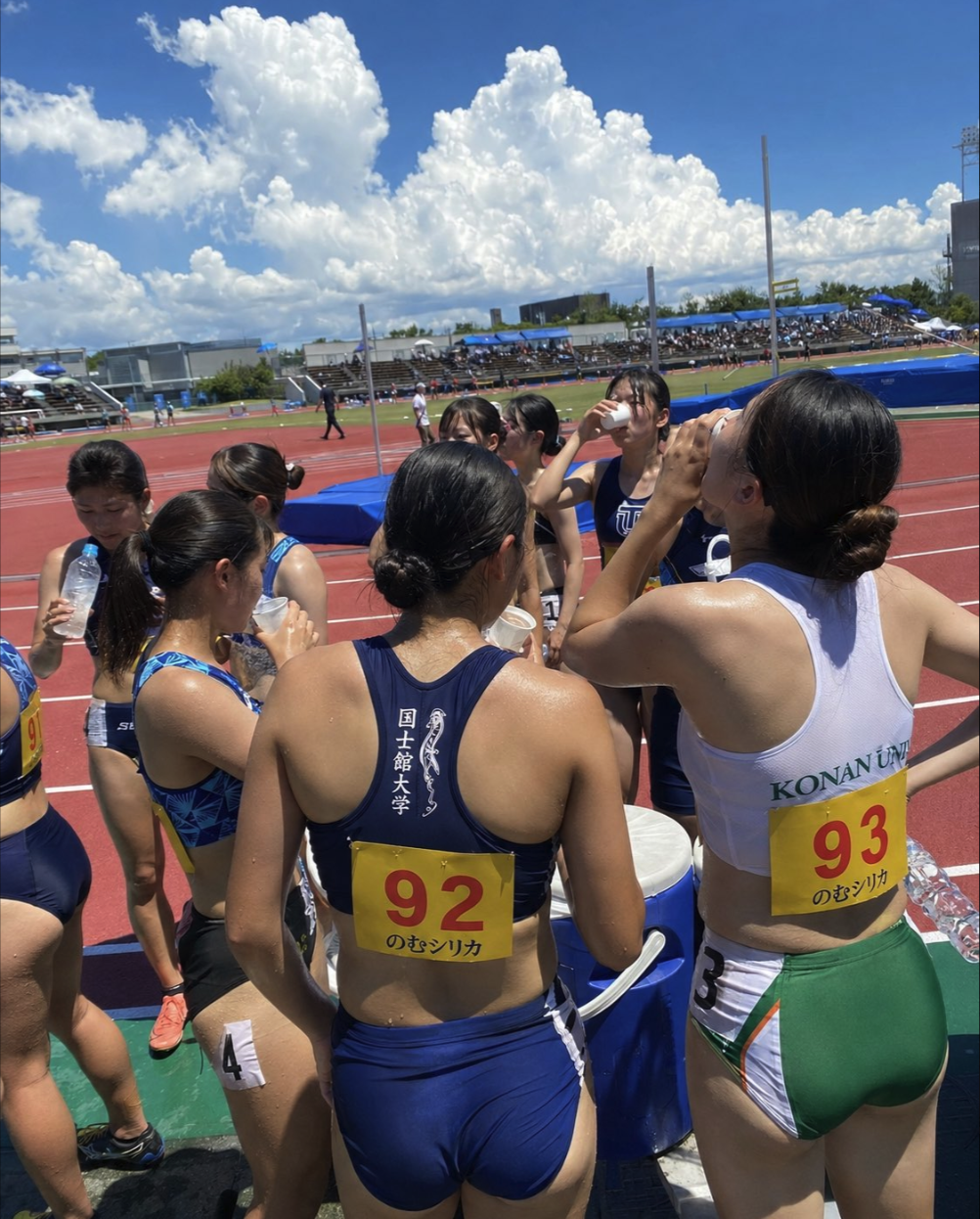 Female athletes gathered on an outdoor running track, drinking water between events.