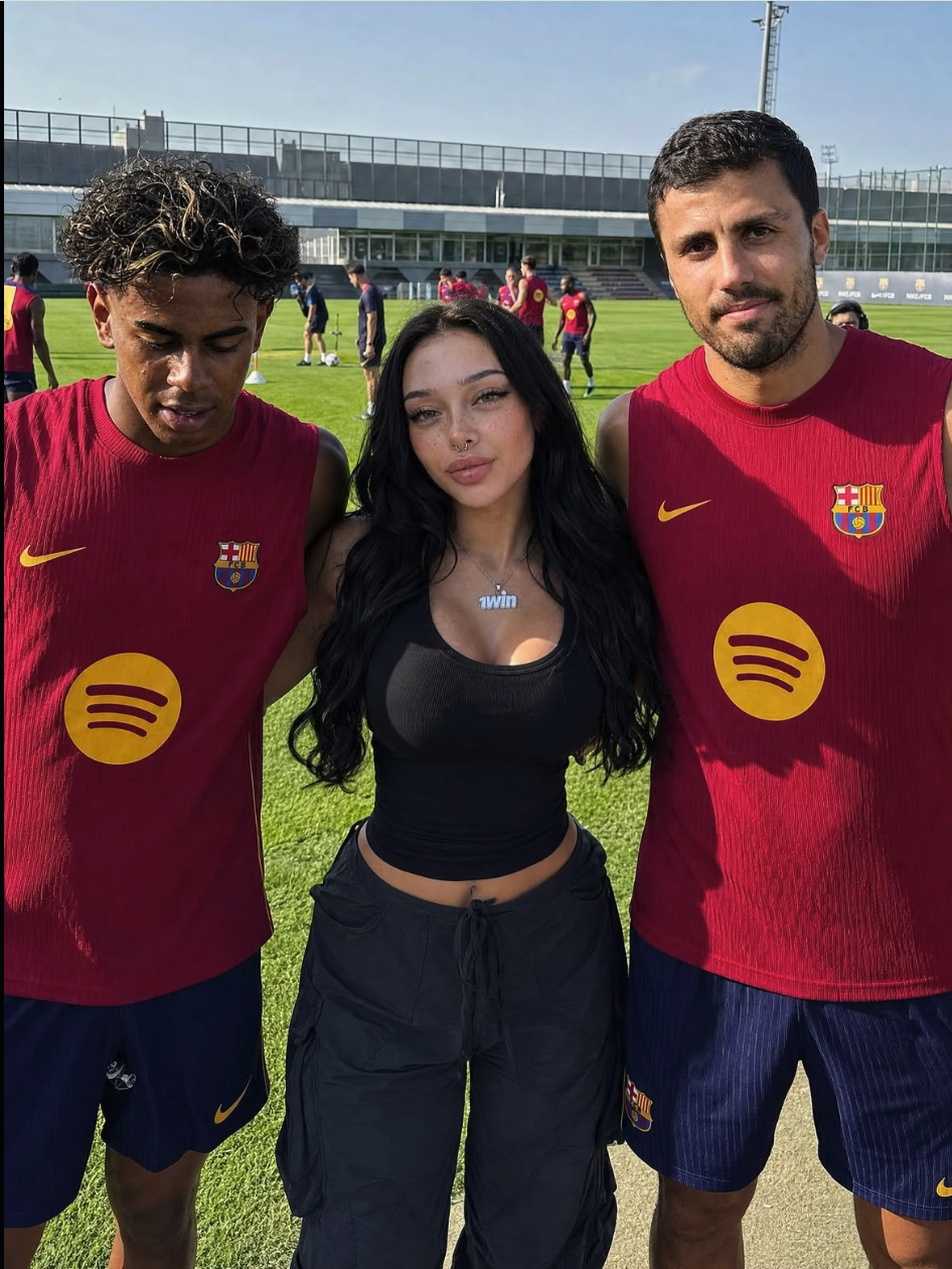 A woman with long dark hair standing between two men in FC Barcelona training kits on a football pitch.