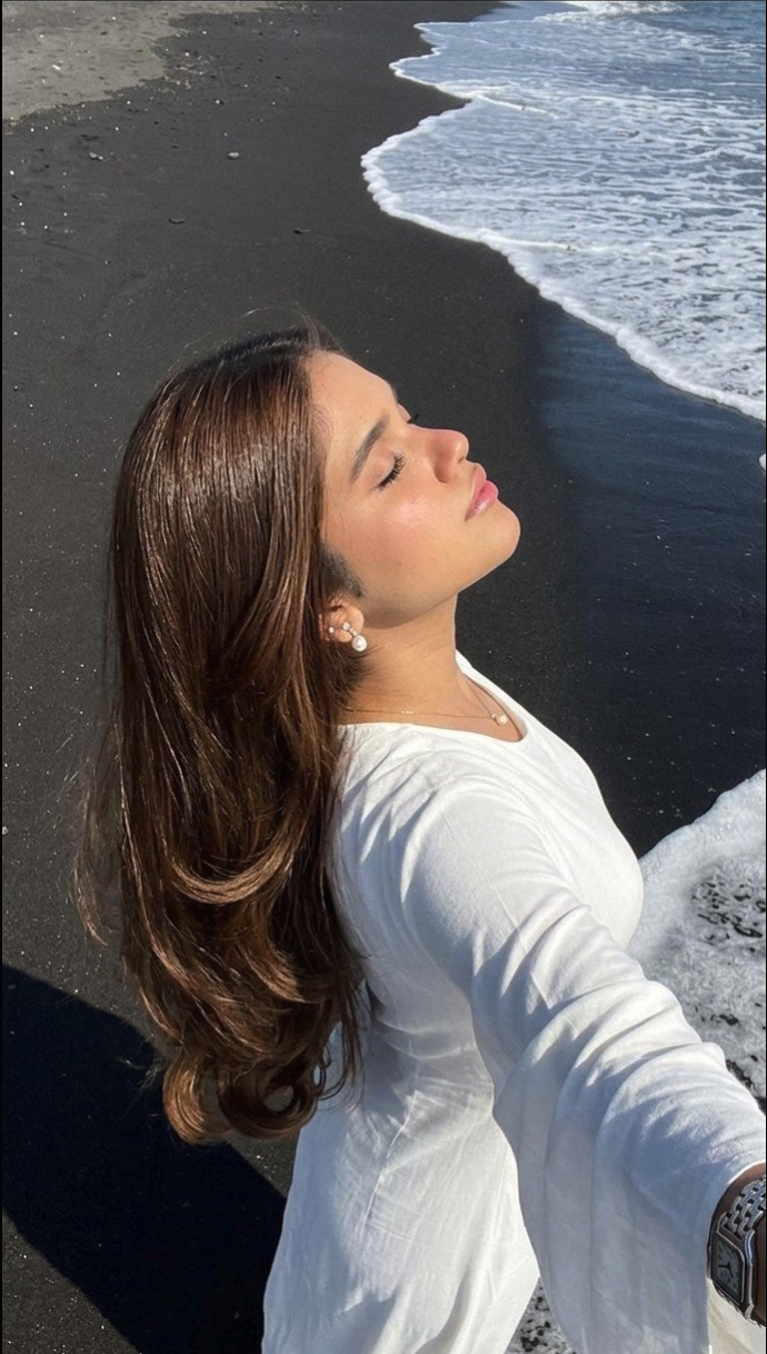 A woman with long brown hair wearing a white long-sleeved shirt, taking a selfie on a dark-sand beach.