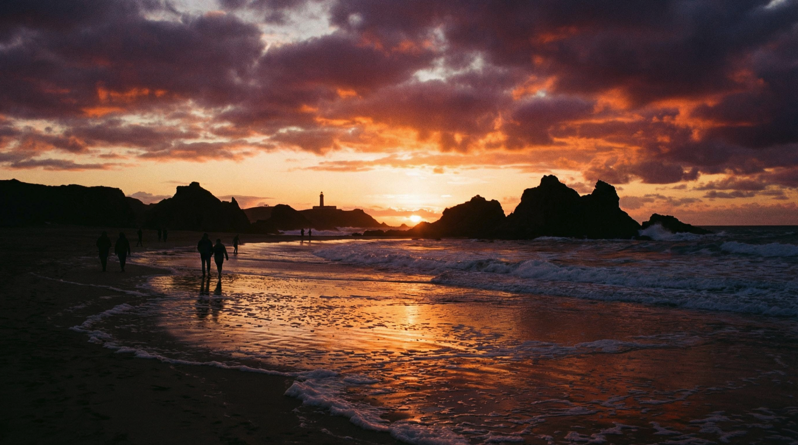 Silhouetted figures walking along a beach at sunset, with rock formations and a distant lighthouse against an orange and purple sky.