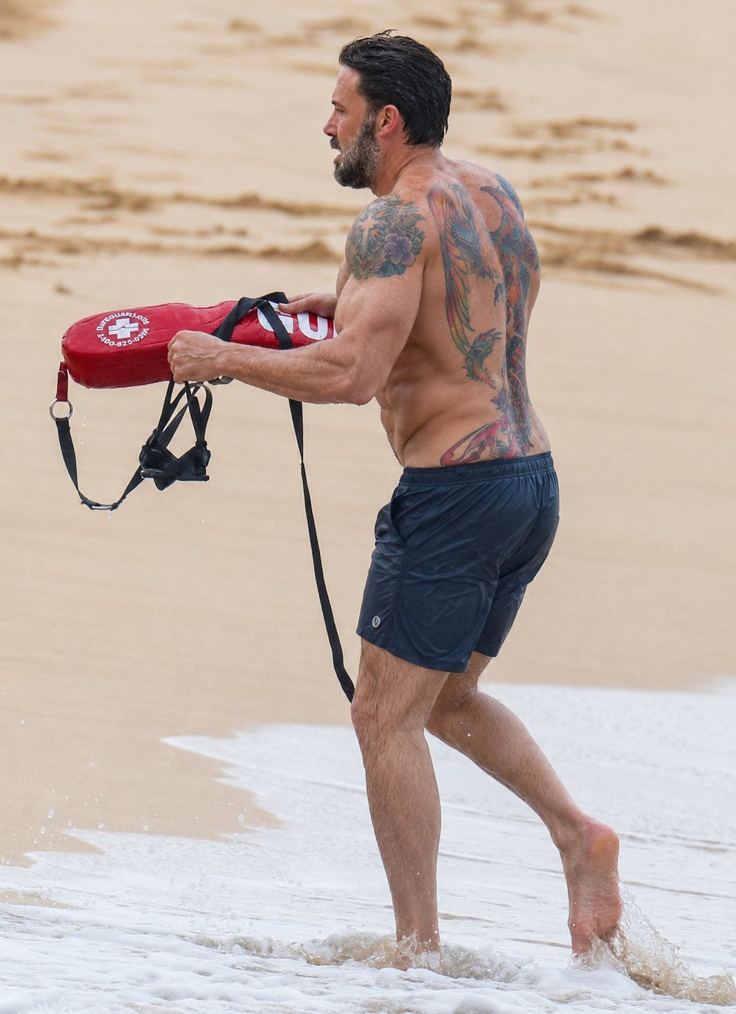 A man resembling Ben Affleck, shirtless with tattoos, carrying a red lifeguard rescue buoy on a sandy beach.