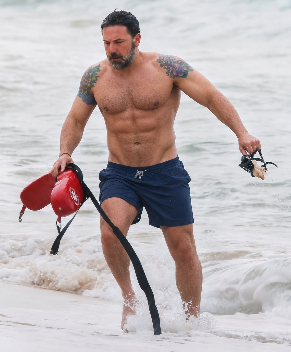 A shirtless man resembling Ben Affleck wading through surf at the beach, holding a red lifeguard rescue tube and a mask.