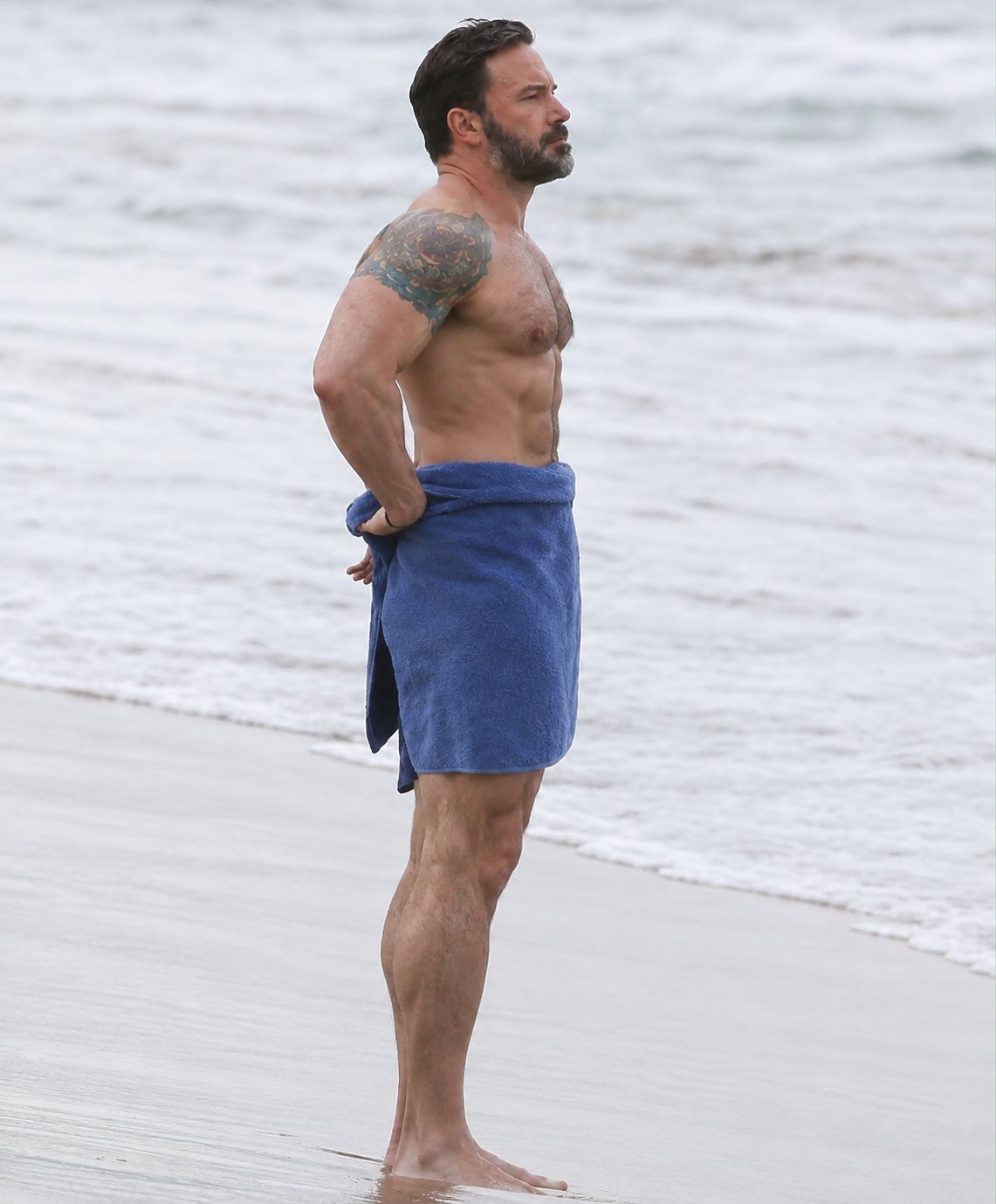 A man resembling Ben Affleck standing on a beach wrapped in a blue towel, facing the water.
