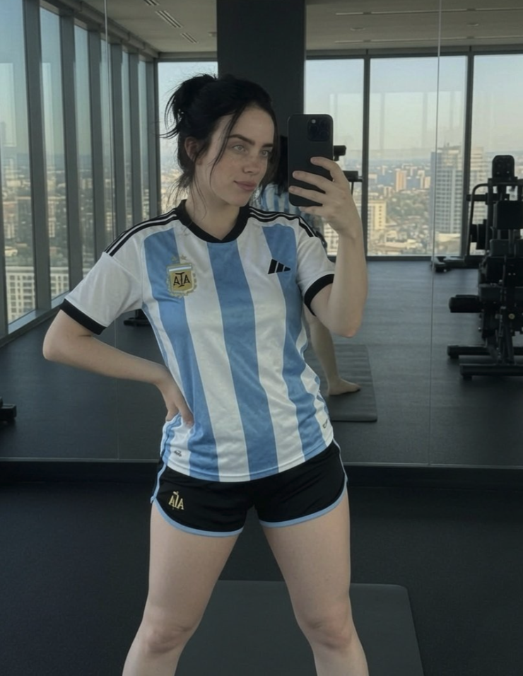 A woman resembling Billie Eilish taking a mirror selfie in an Argentina national football jersey and black shorts inside a gym.