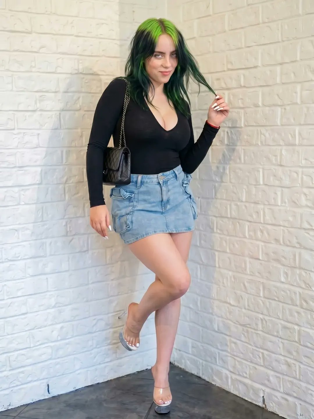 A woman resembling Billie Eilish with green-streaked hair, wearing a black long-sleeve top and denim mini skirt, standing against a white brick wall.