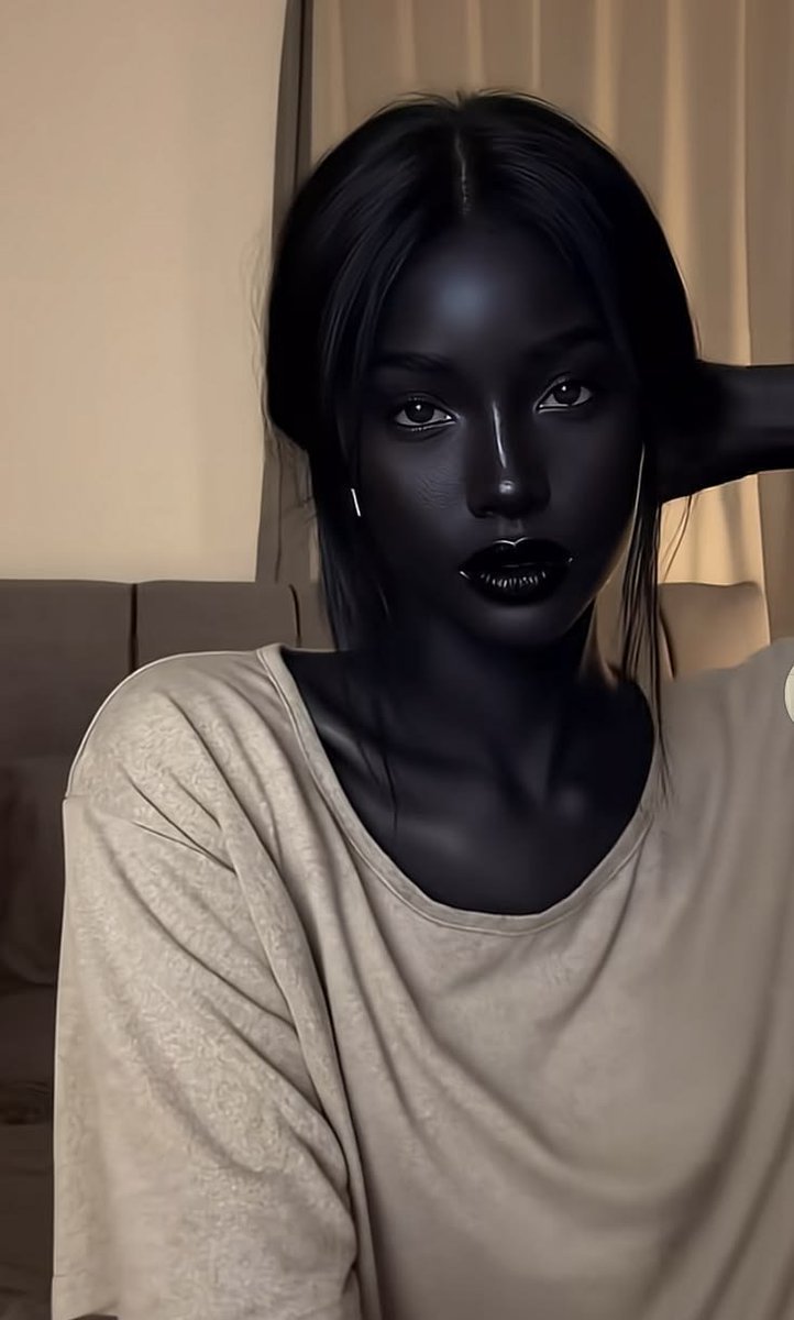 A woman appearing in a selfie-style portrait with entirely black-painted skin, dark glossy lips, and long dark hair, seated indoors on a beige sofa.