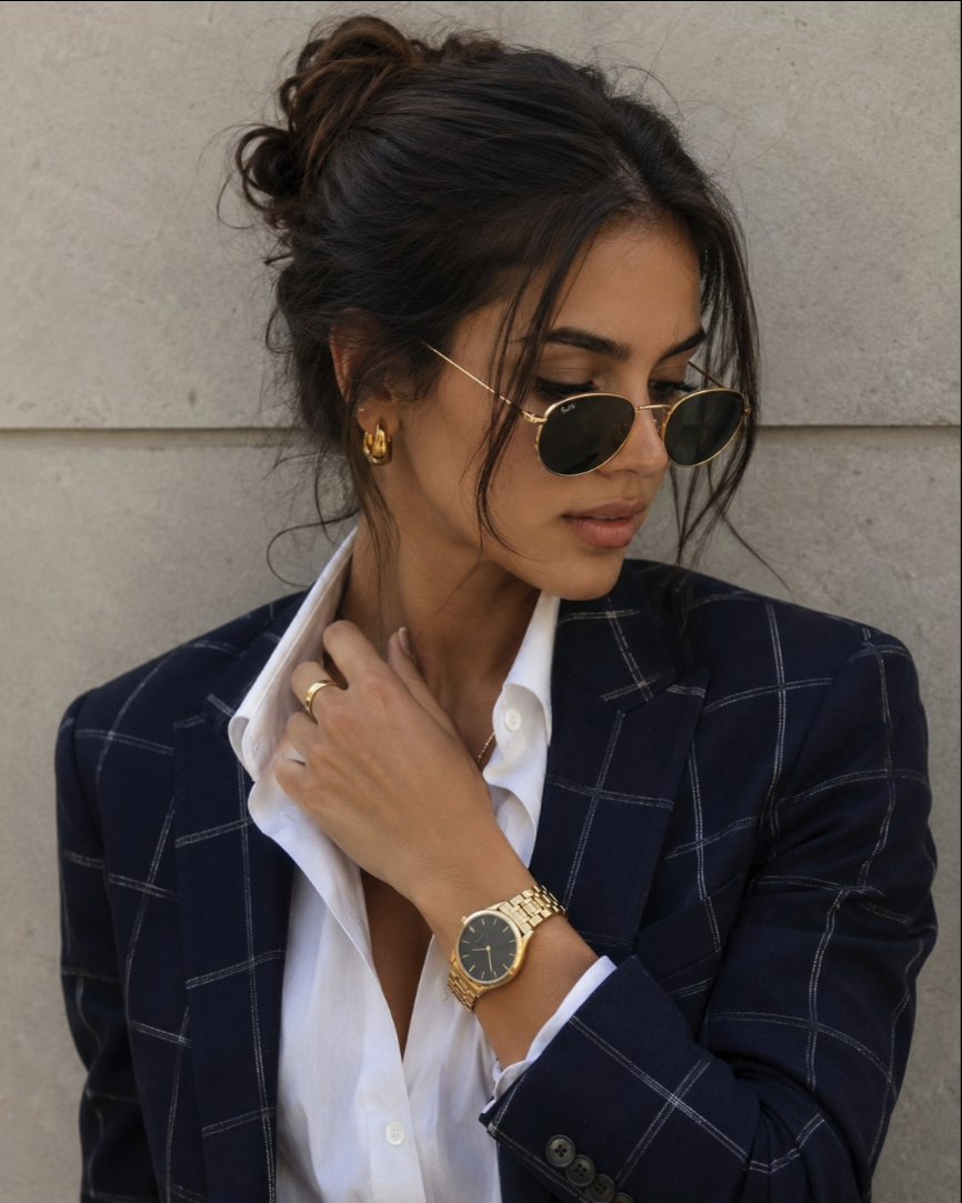 A woman wearing a navy checked blazer, white shirt, and gold sunglasses.