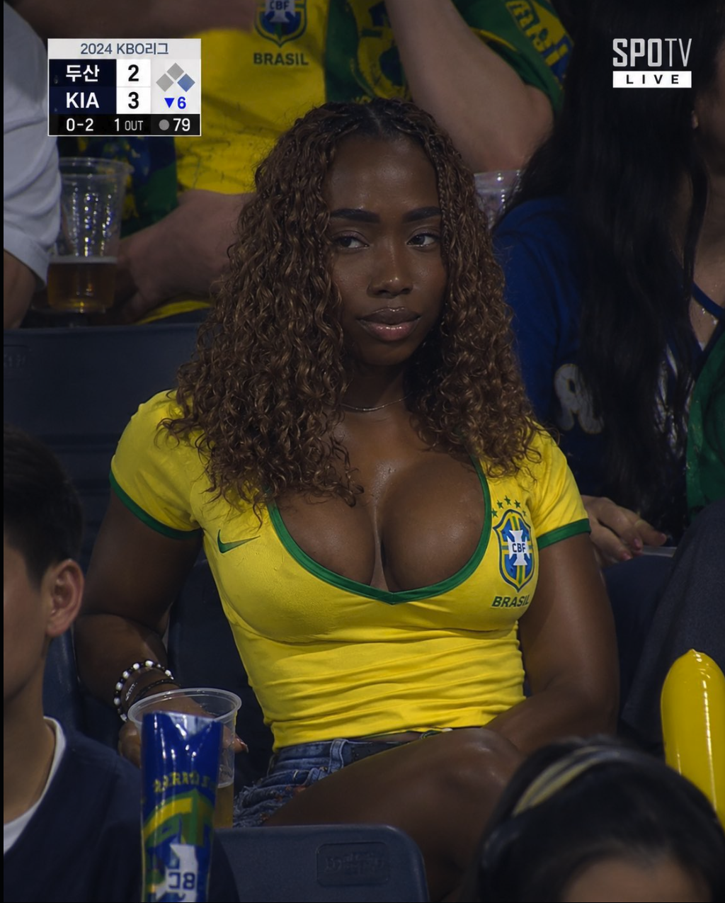 A woman wearing a yellow Brazil national team jersey seated in stadium stands, with a KBO League scoreboard visible in the background.