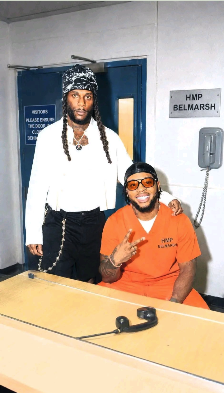 A man resembling Burna Boy seated in an orange HMP Belmarsh jumpsuit beside a standing man with long braids in a prison visitation room.
