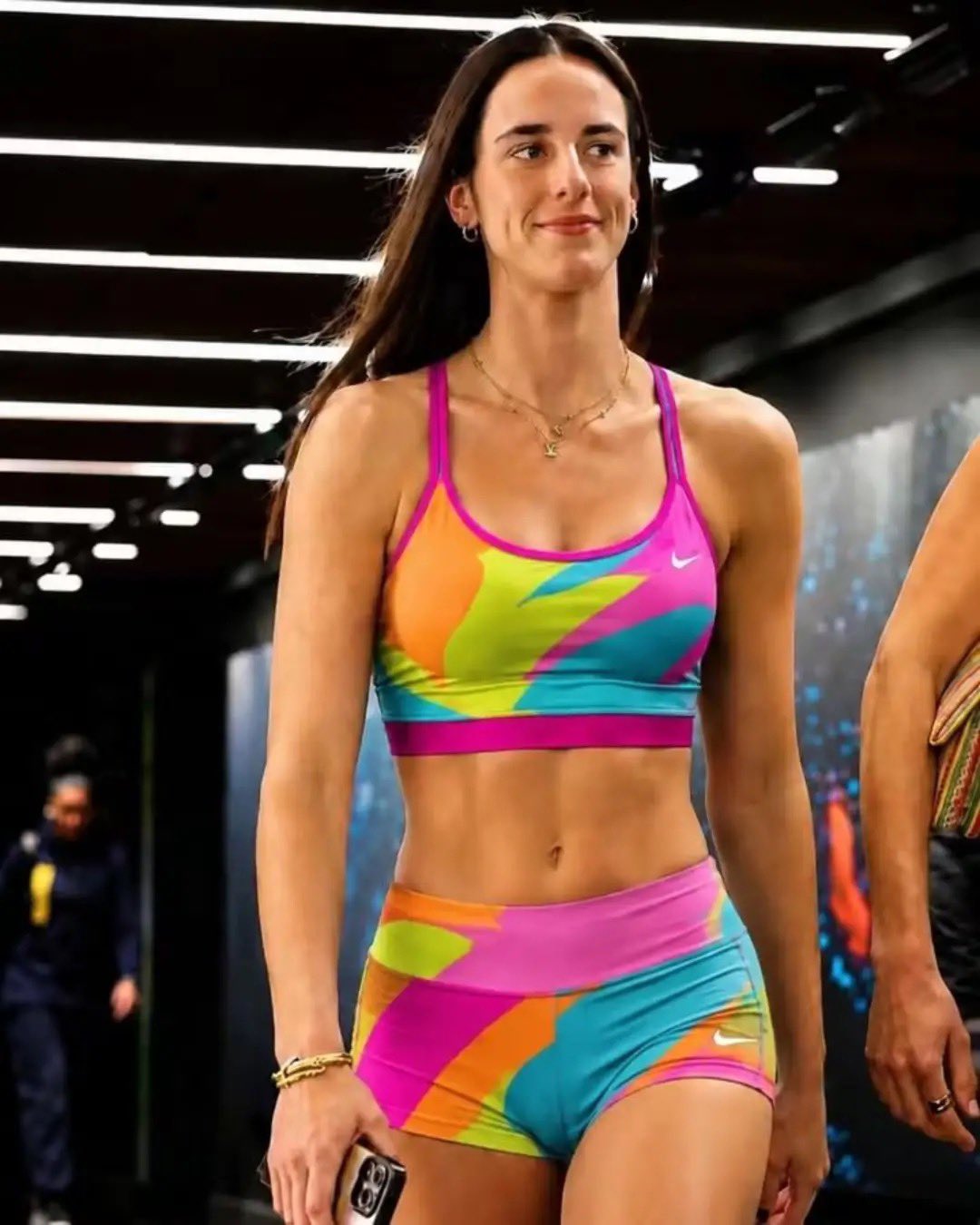 A woman resembling Caitlin Clark walking through an arena tunnel wearing a colorful Nike sports bra and matching shorts.
