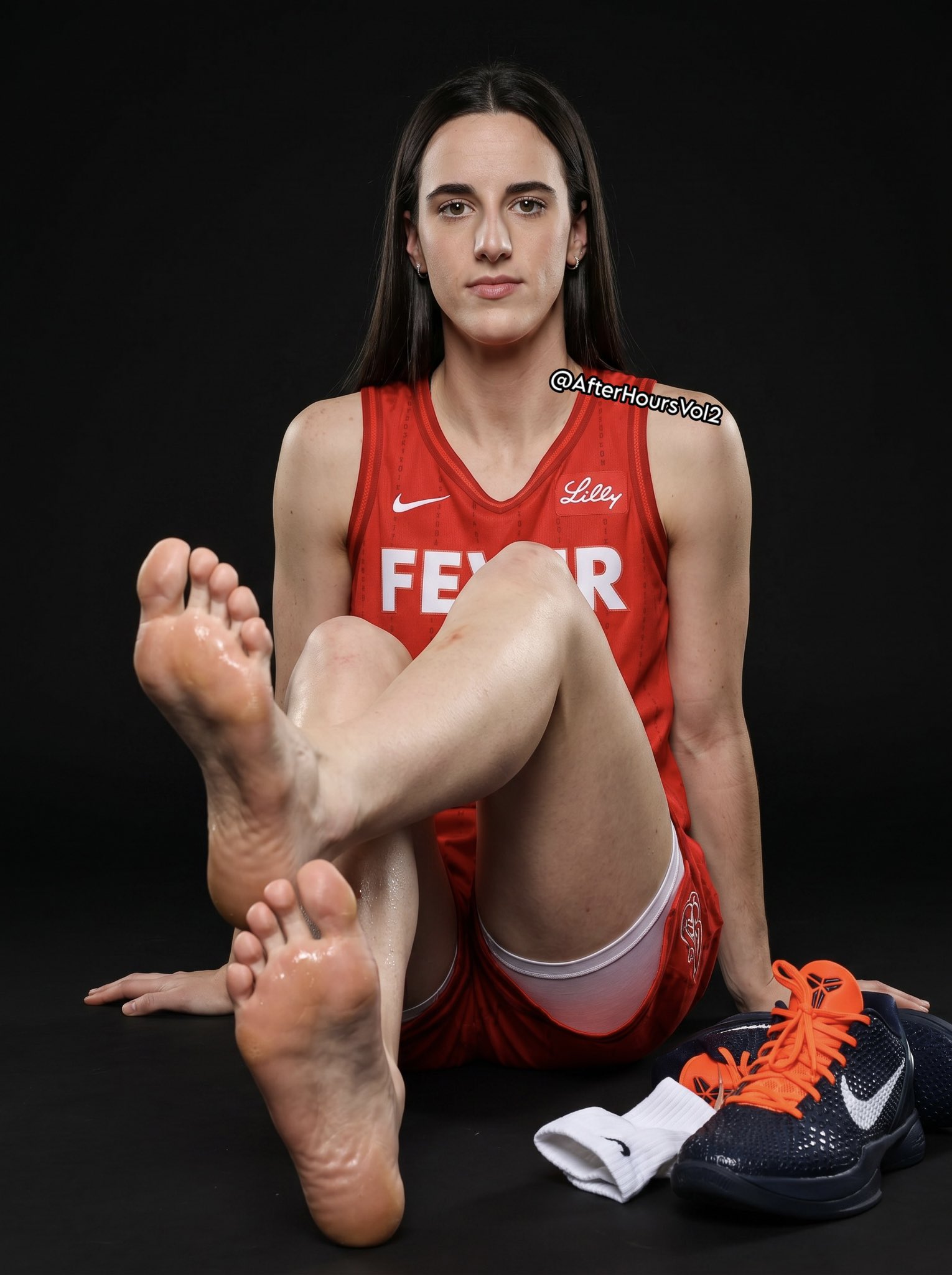 A woman resembling Caitlin Clark seated on a dark studio floor in a red Indiana Fever jersey, with bare feet extended toward the camera and a pair of basketball shoes beside her.