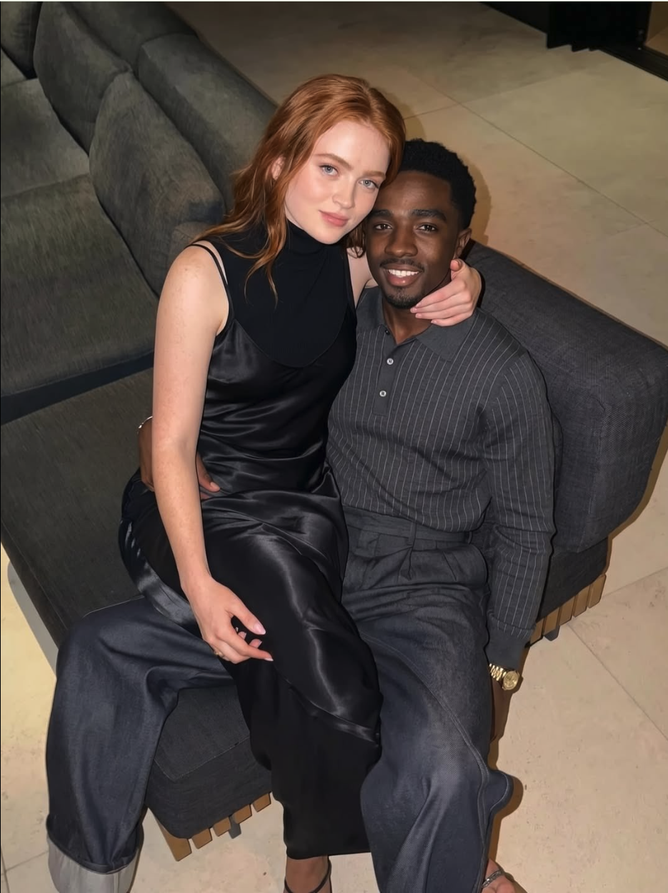 A woman with auburn hair resembling Sadie Sink sits on the lap of a man resembling Caleb McLaughlin on a gray couch.