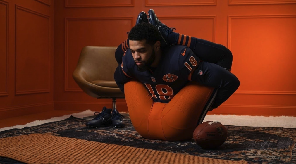 A man resembling Caleb Williams posing in a Chicago Bears #18 uniform on an orange cushion inside an orange-walled studio.