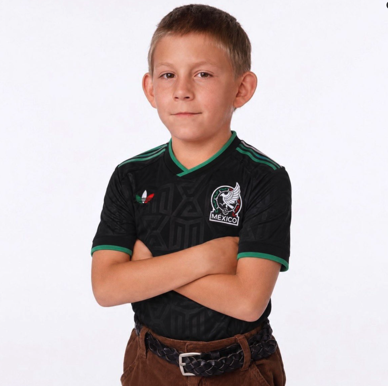 A young boy with short brown hair standing with arms crossed against a white background, wearing a Mexico national team soccer jersey.