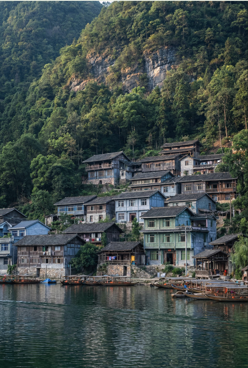 A hillside settlement of traditional wooden houses with dark tiled roofs above a calm river, backed by a forested cliff.