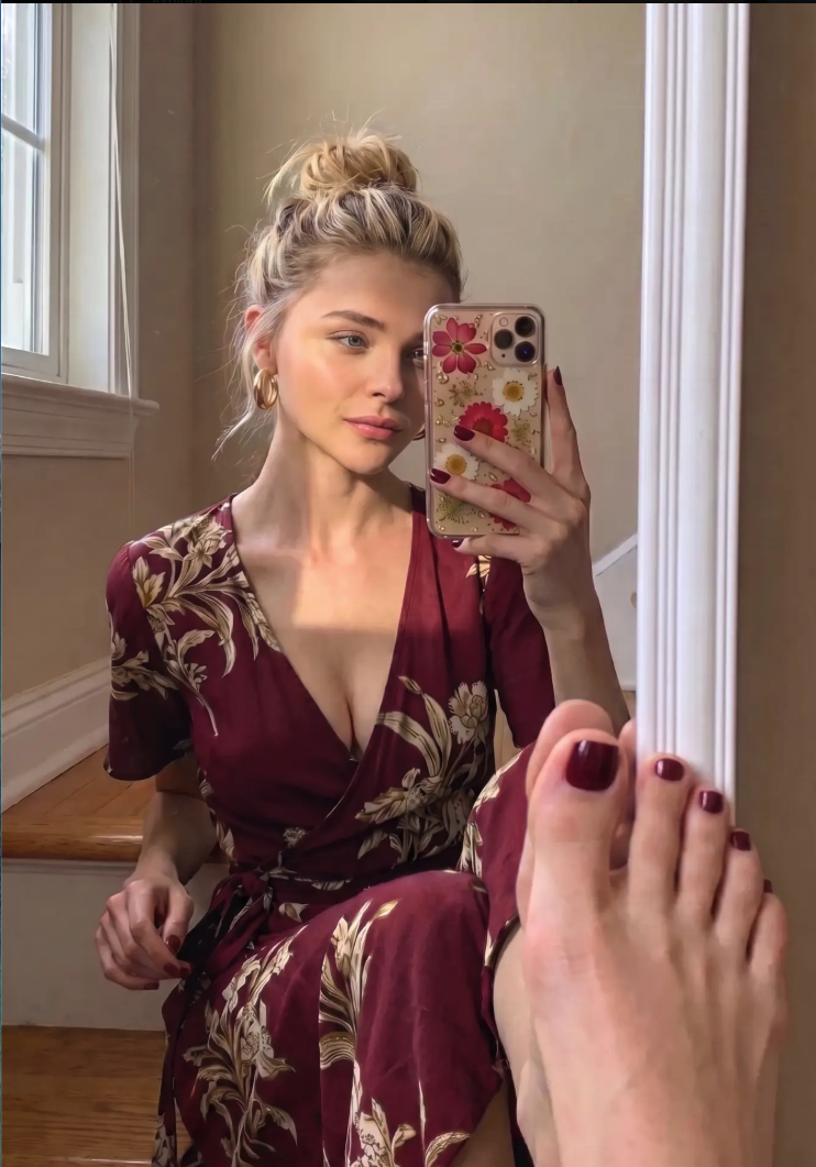 A woman resembling Chloë Grace Moretz taking a mirror selfie while wearing a burgundy floral wrap dress.