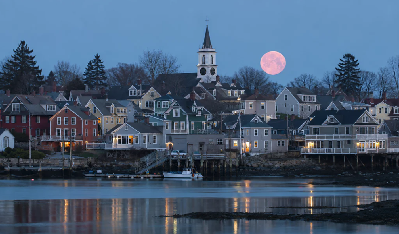 A coastal New England town at twilight featuring a white church steeple, colorful houses, and an unusually large pink full moon over the harbor.