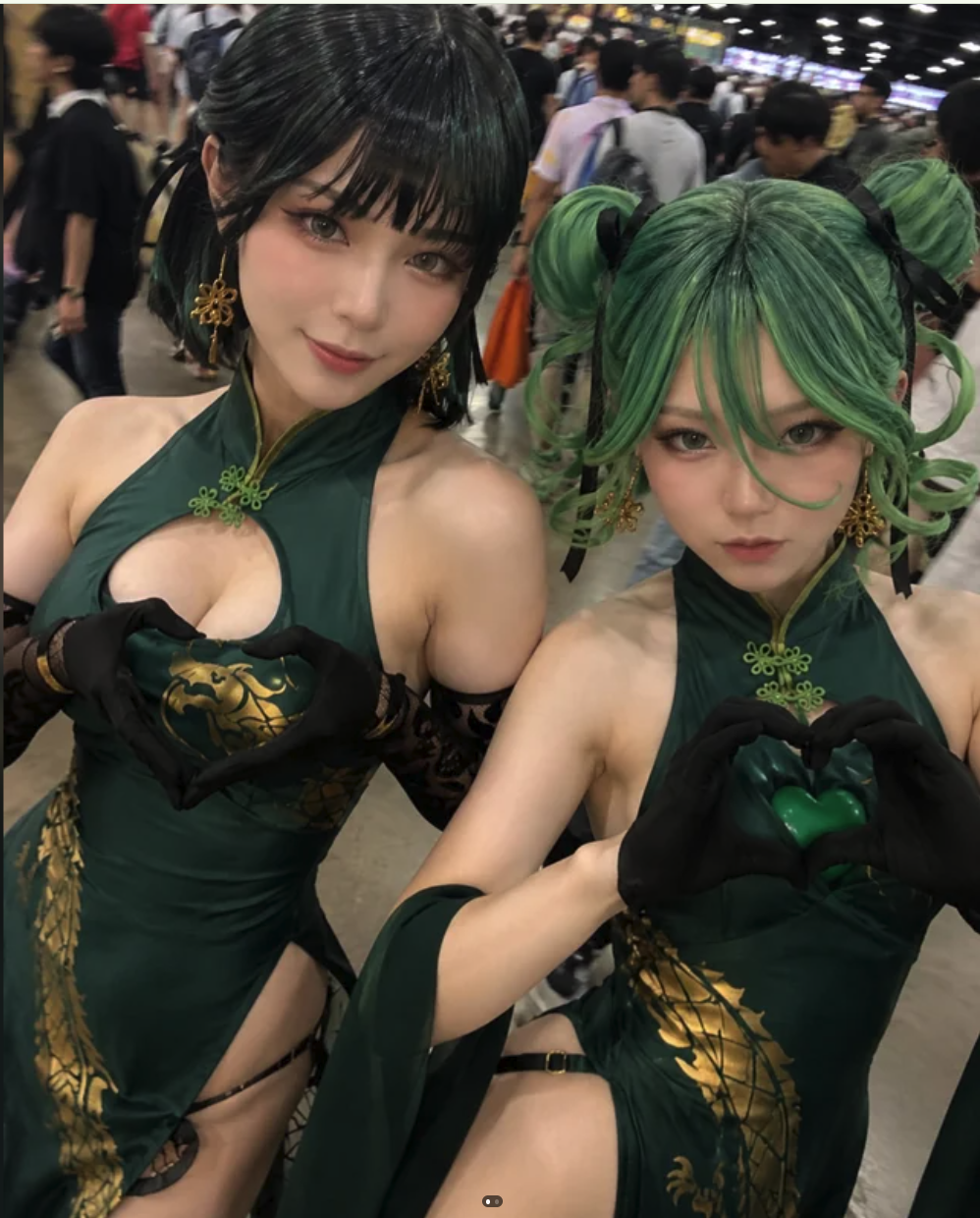 Two women in green Chinese-style dresses forming heart shapes with their hands at a convention hall.