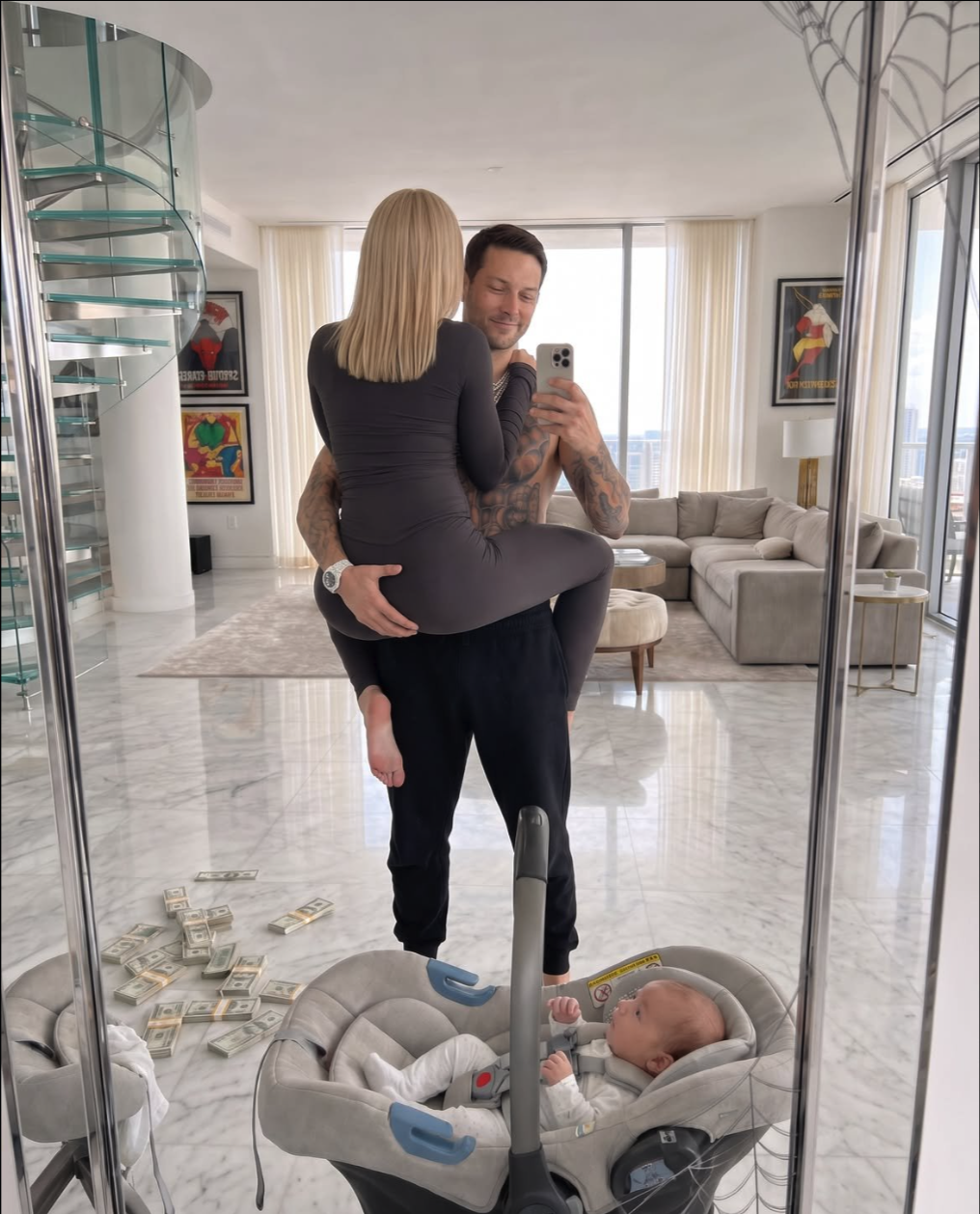 A man holding a woman while taking a mirror selfie in a modern apartment, with an infant in a car seat and scattered cash on the marble floor.