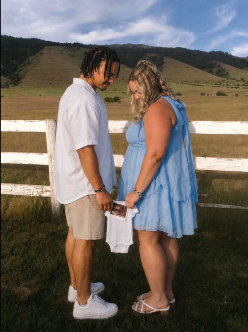 A man and woman standing in a grassy field, looking down at a baby onesie and an ultrasound print they are holding between them.