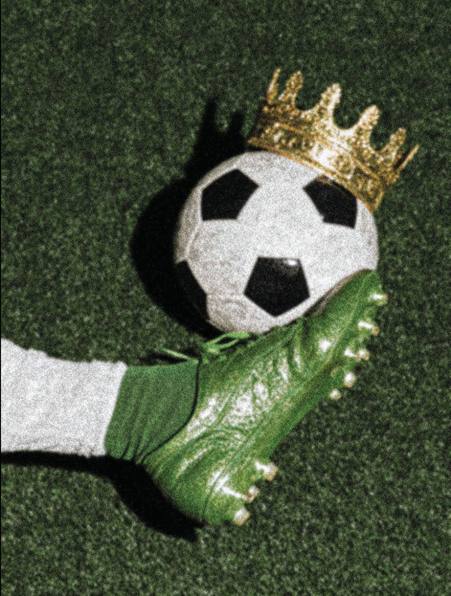 A soccer ball topped with a gold crown, held by a green and white cleated foot against a grassy background.
