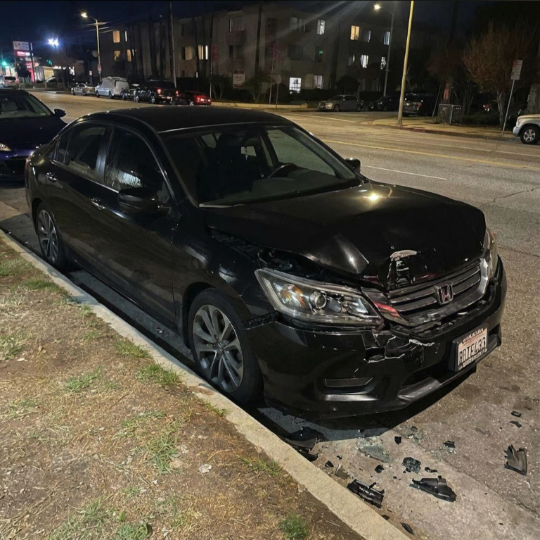 A damaged black Honda sedan parked on a curb at night, with a broken headlight and debris on the ground.