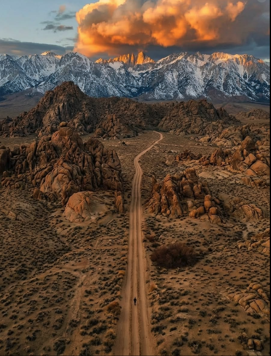 A dirt road winding through a rocky desert toward snow-capped mountains lit by a golden sunset, with a small lone figure walking along the road.