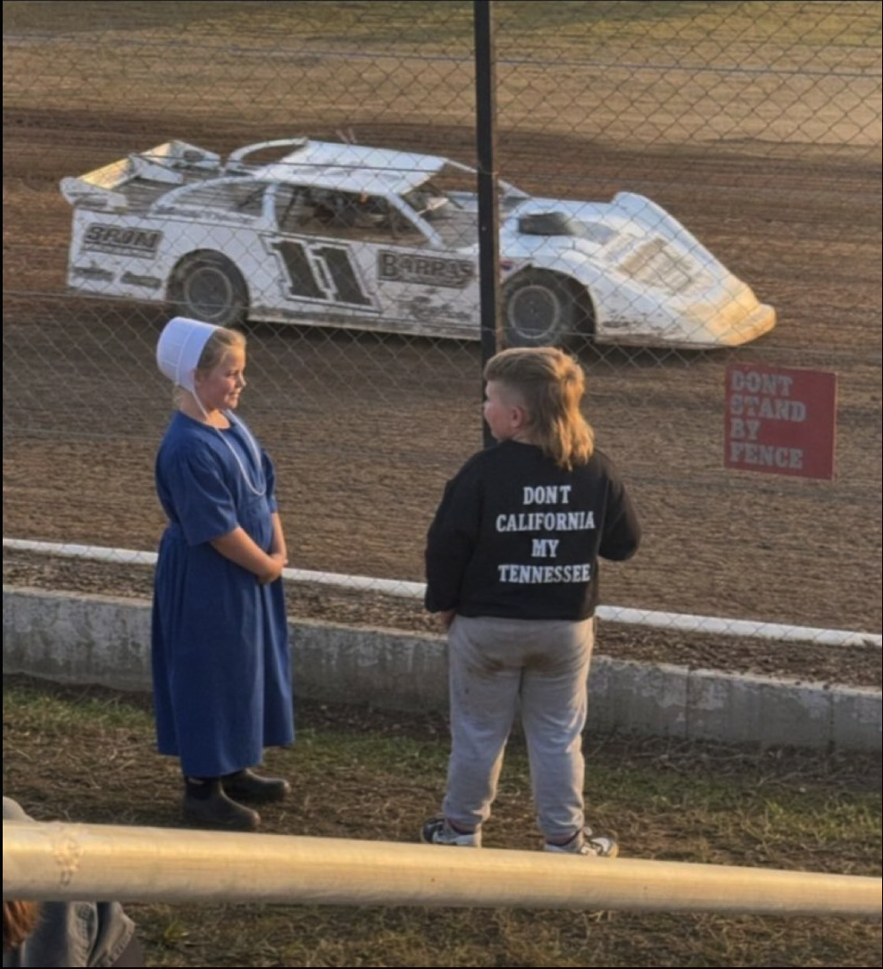 Two children standing near a chain-link fence in front of a dirt racetrack with a wrecked race car.