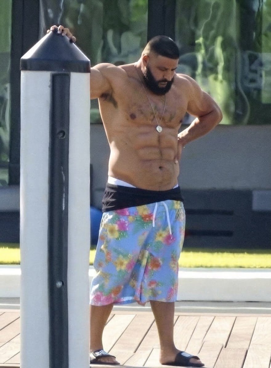 A man resembling DJ Khaled standing shirtless in floral swim shorts beside a post on an outdoor deck with six pack abs.