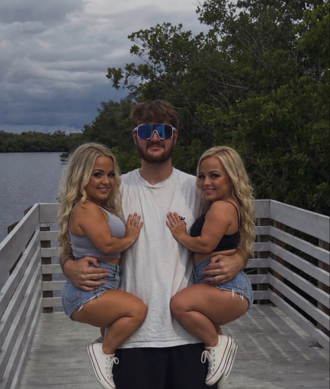 A man in sunglasses flanked by two blonde women posing together on a wooden dock beside a lake.