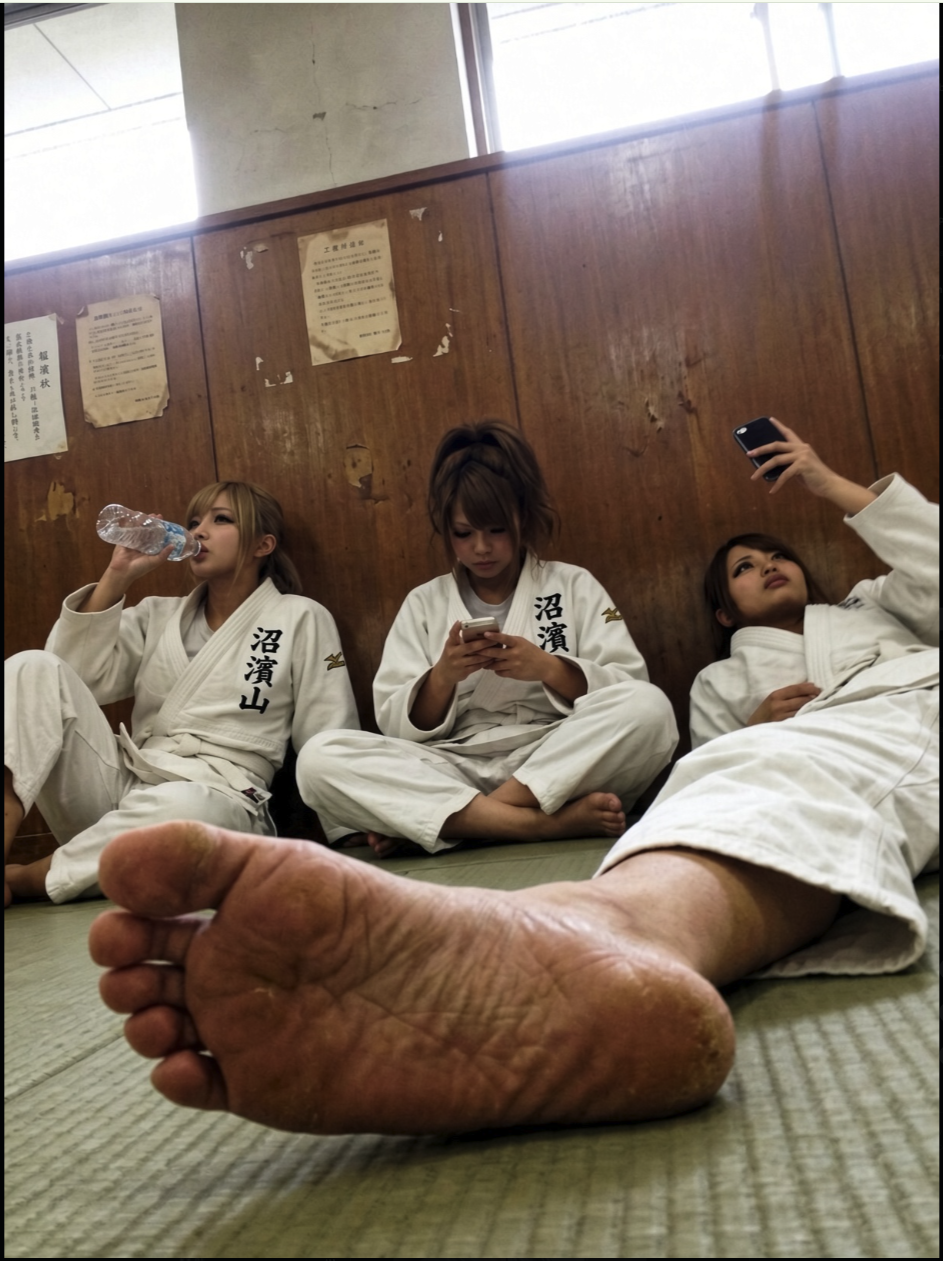Three women in white martial arts gis seated on tatami mats in a dojo, with a bare foot in the foreground.