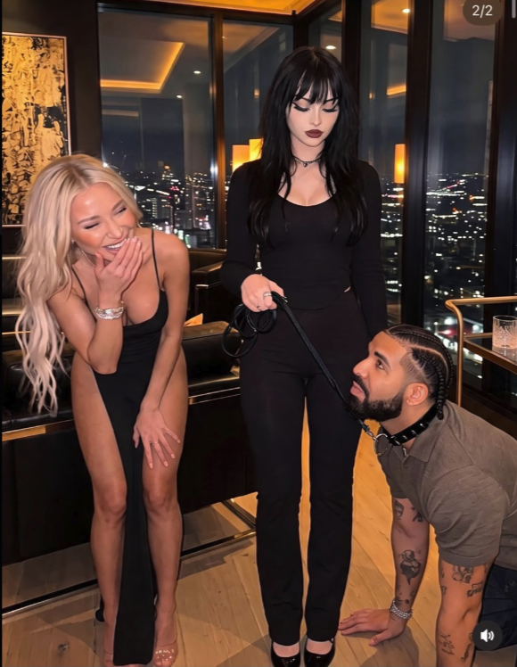 A man resembling Drake kneeling on a leash held by a dark-haired woman, while a blonde woman laughs nearby, in an upscale high-rise interior at night.