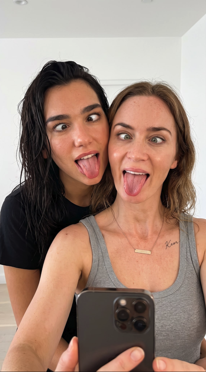 Two women resembling Emily Blunt and Dua Lipa taking a close selfie together, both sticking their tongues out.