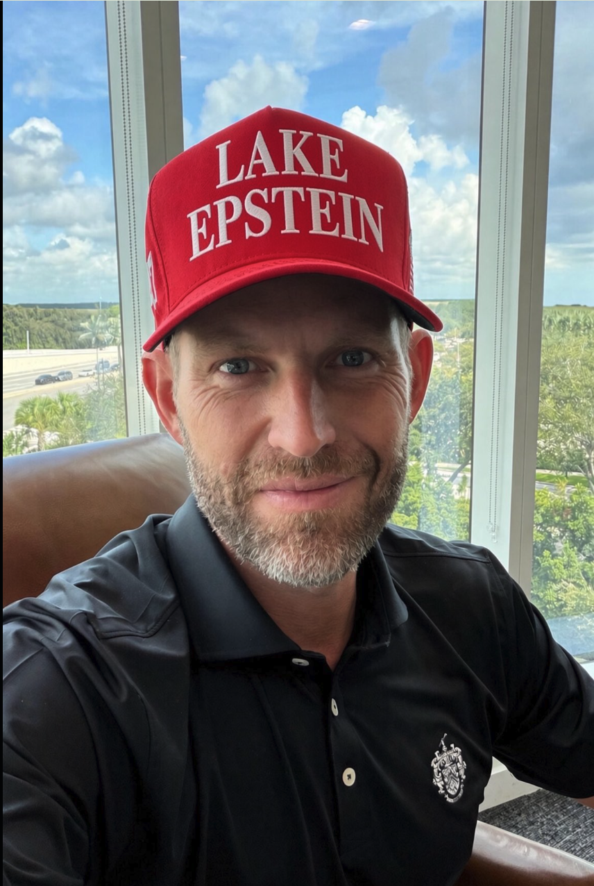 A man resembling Eric Trump wearing a red 'LAKE EPSTEIN' cap and a black polo shirt by a window.
