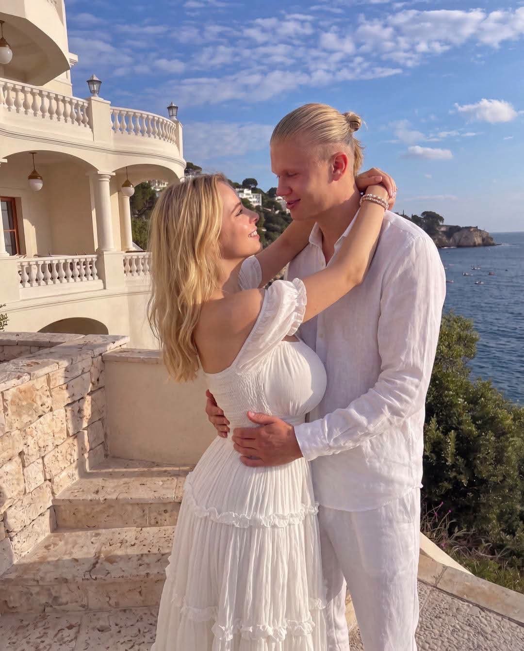 A man resembling Erling Haaland and a blonde woman embracing on a sunlit coastal terrace in white clothing.