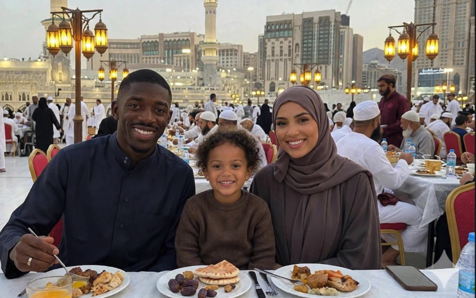 A man, a woman wearing a hijab, and a young child seated at a dining table with plates of food, with the Grand Mosque in Mecca in the background.