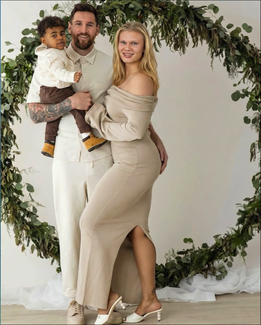 A man holding a young child beside a pregnant woman in an off-shoulder dress, posed in front of a floral greenery arch.