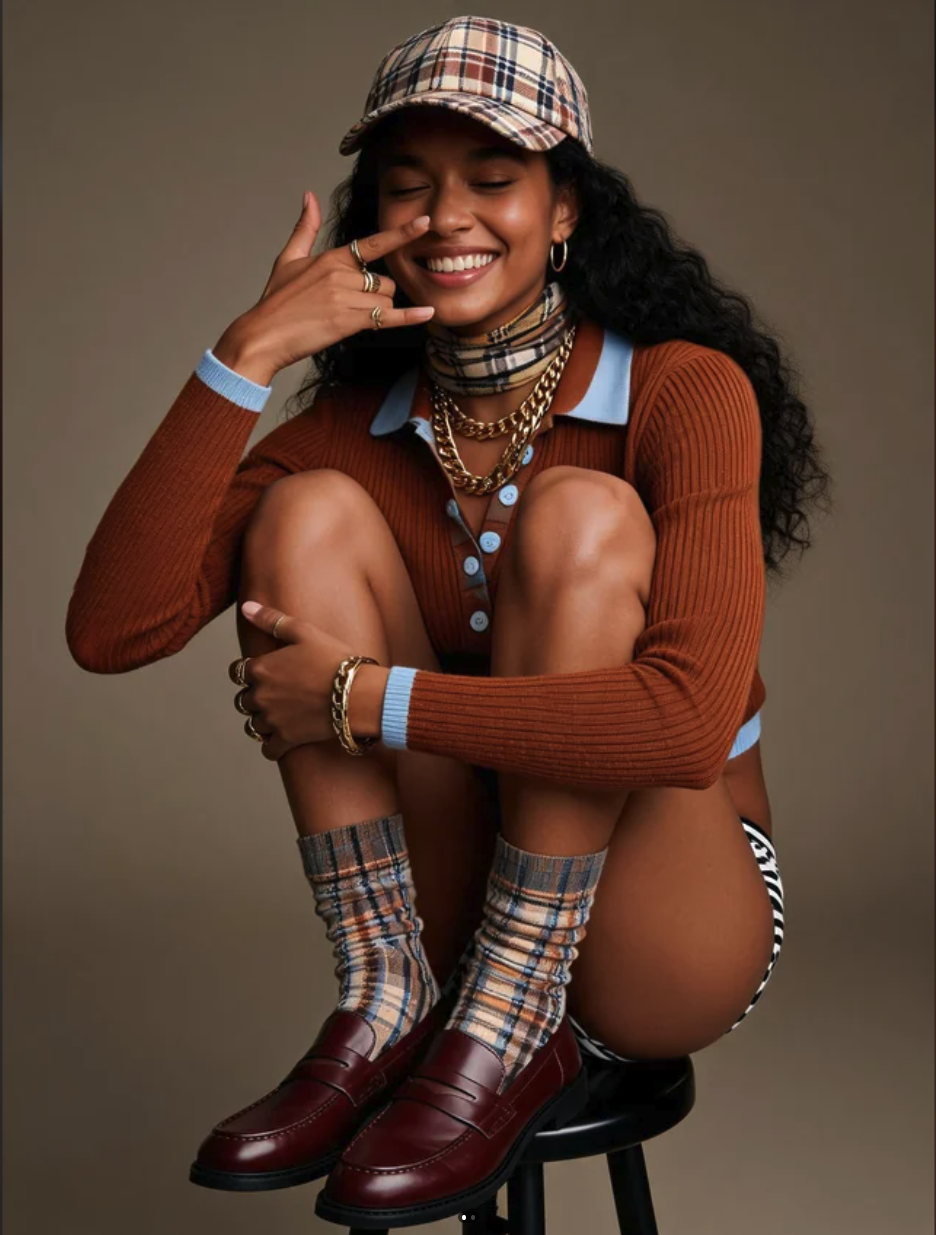 A woman smiling with her eyes closed, wearing a plaid cap, layered gold jewelry, an orange cardigan, plaid socks, and burgundy loafers.