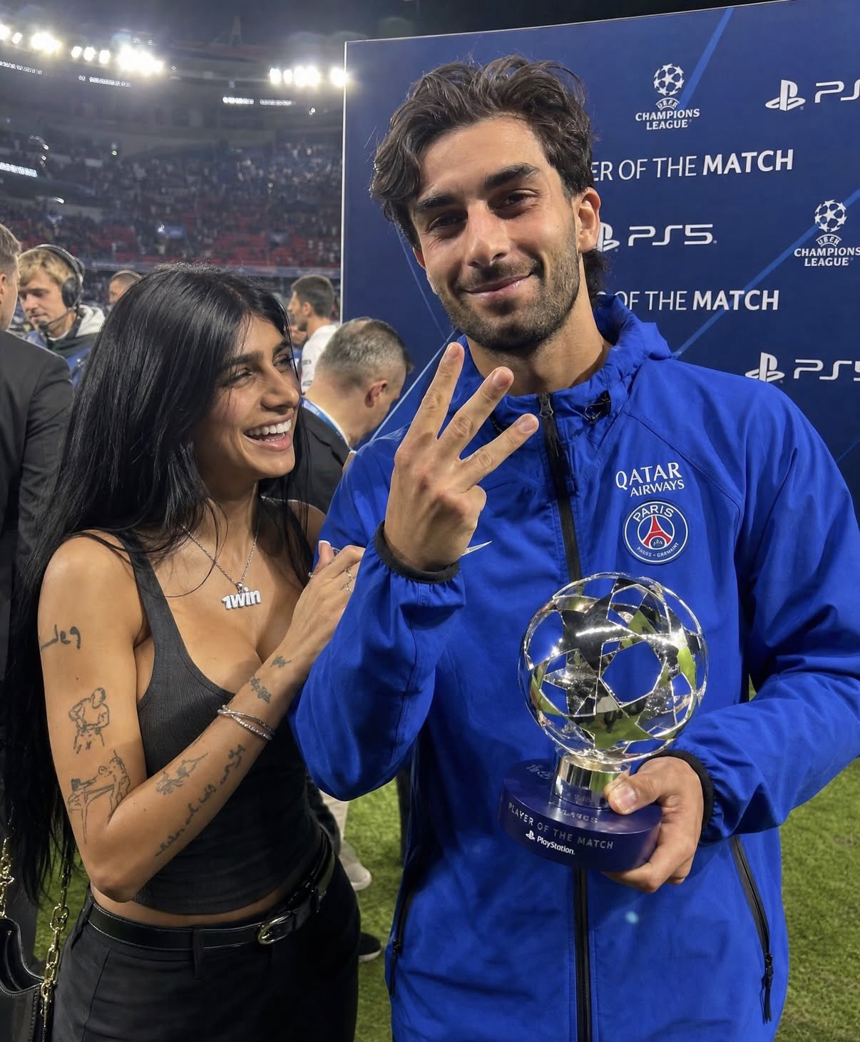 A man resembling Ferran Torres in a blue PSG jacket holding a Champions League Player of the Match trophy beside a woman resembling Mia Khalifa.