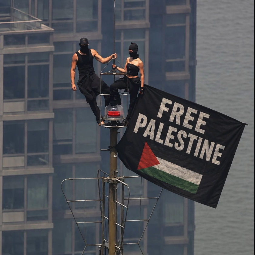 Two masked figures in black clothing on a ship's mast holding a large 'Free Palestine' flag.